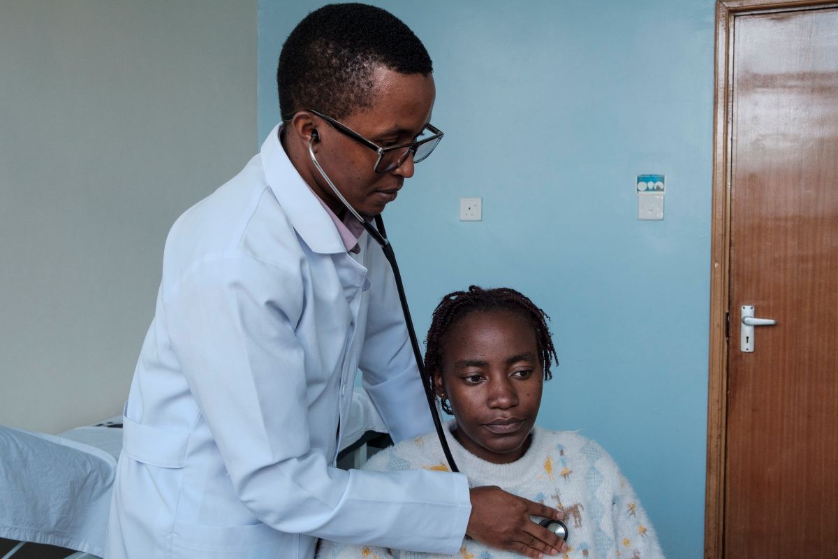 An African (male) doctor listens with a stethoscope to a female patient's heart (seated, robed)