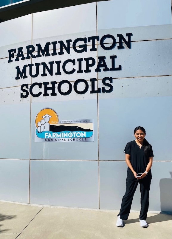A young girl stands outside a sign reading "Farmington Municipal Schools."