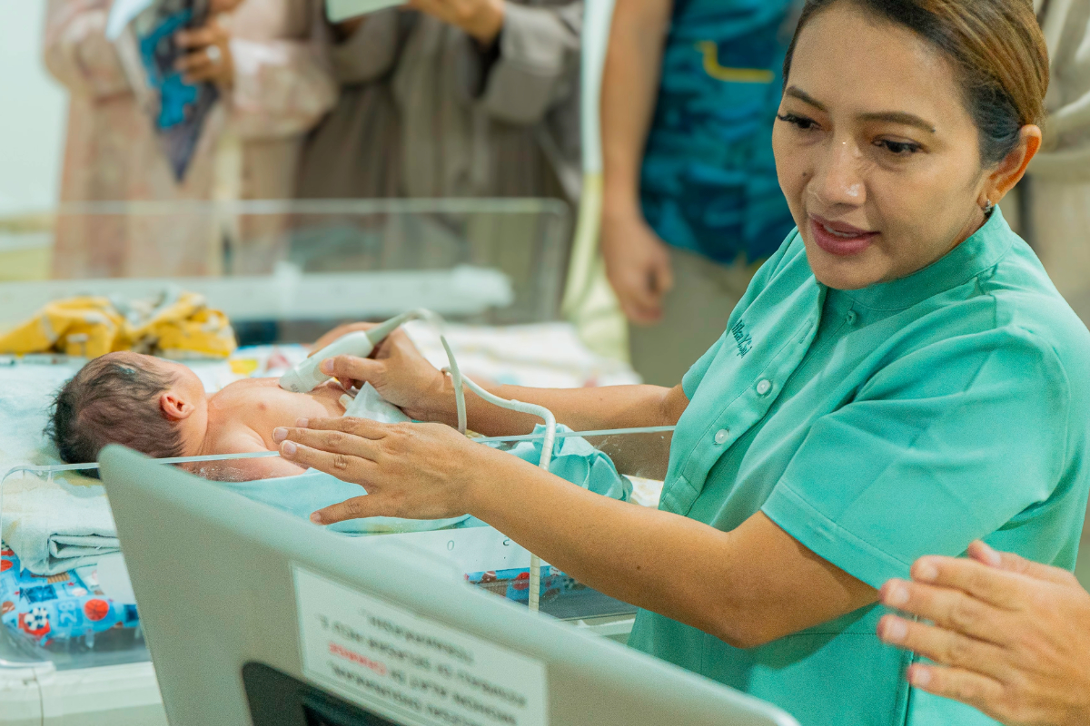 A female doctor looks at an ultrasound monitor. A newborn sleeps in a hospital crib next to her.