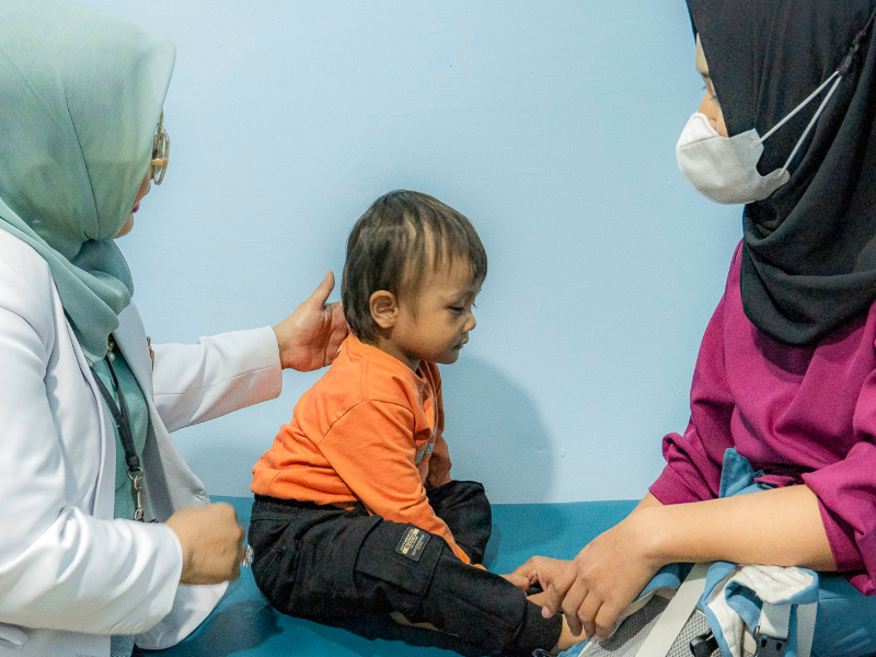 A baby boy sits between his mom and his doctor on an exam table in Indonesia.
