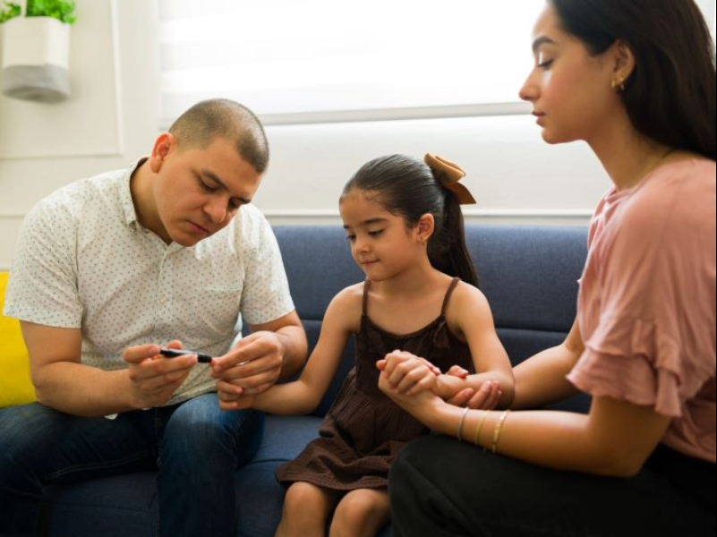Adobe stock, a young girl sits between her parents on a couch. Her dad pricks her finger.
