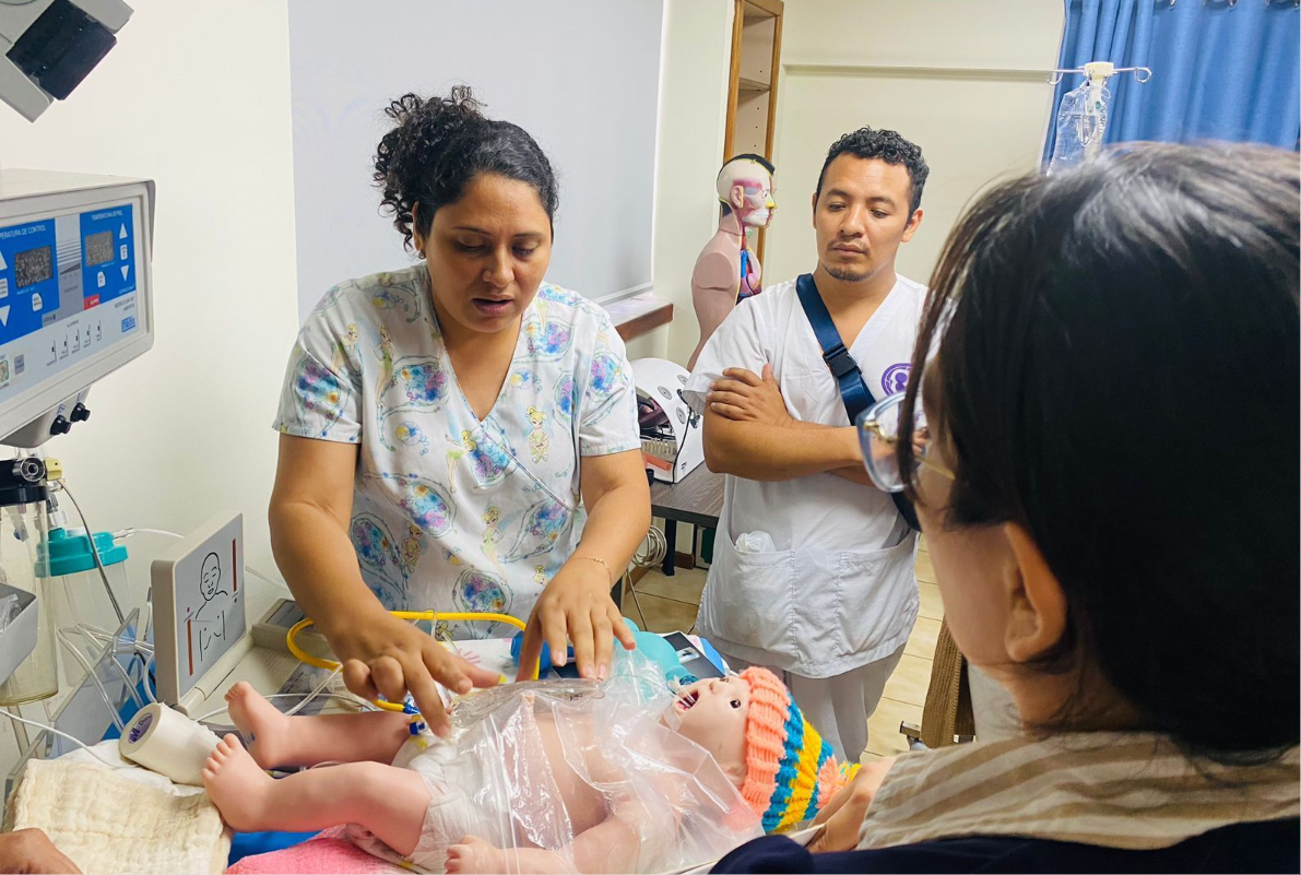 A nurse stands over a newborn; two adults stand by.