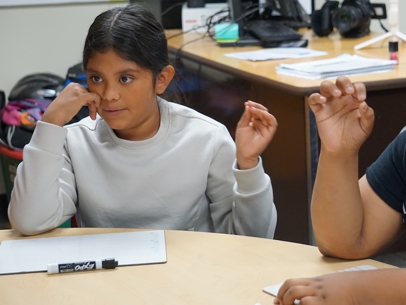 A fifth-grade Hispanic girl listens attentively to her teacher and follows along with the phonics lesson by using her hand to demonstrate sounds.