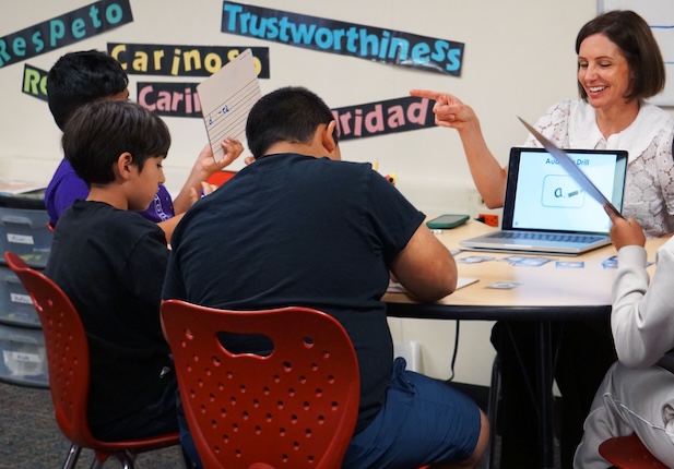 A group of fifth grade boys look at their papers in front of them. Their teacher smiles and points