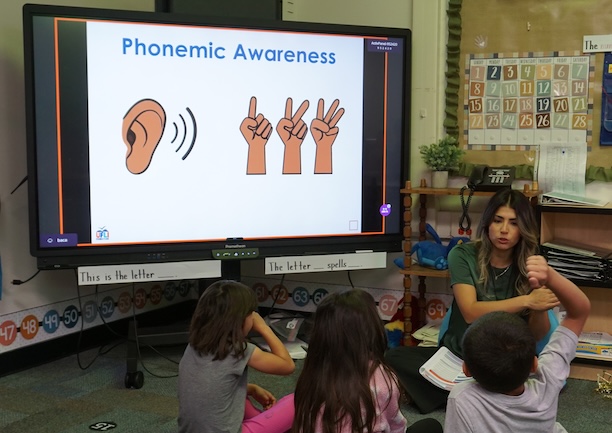 A group of kindergarten students sit on a carpet in front of their teacher. A large screen reads "Phonemic Awareness"