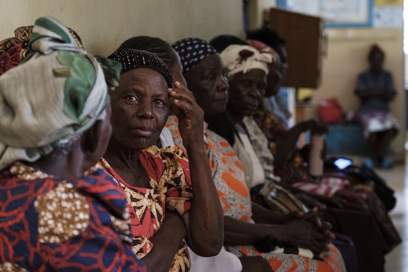 A older woman, seated in a waiting room, looks at the camera and touches her head.