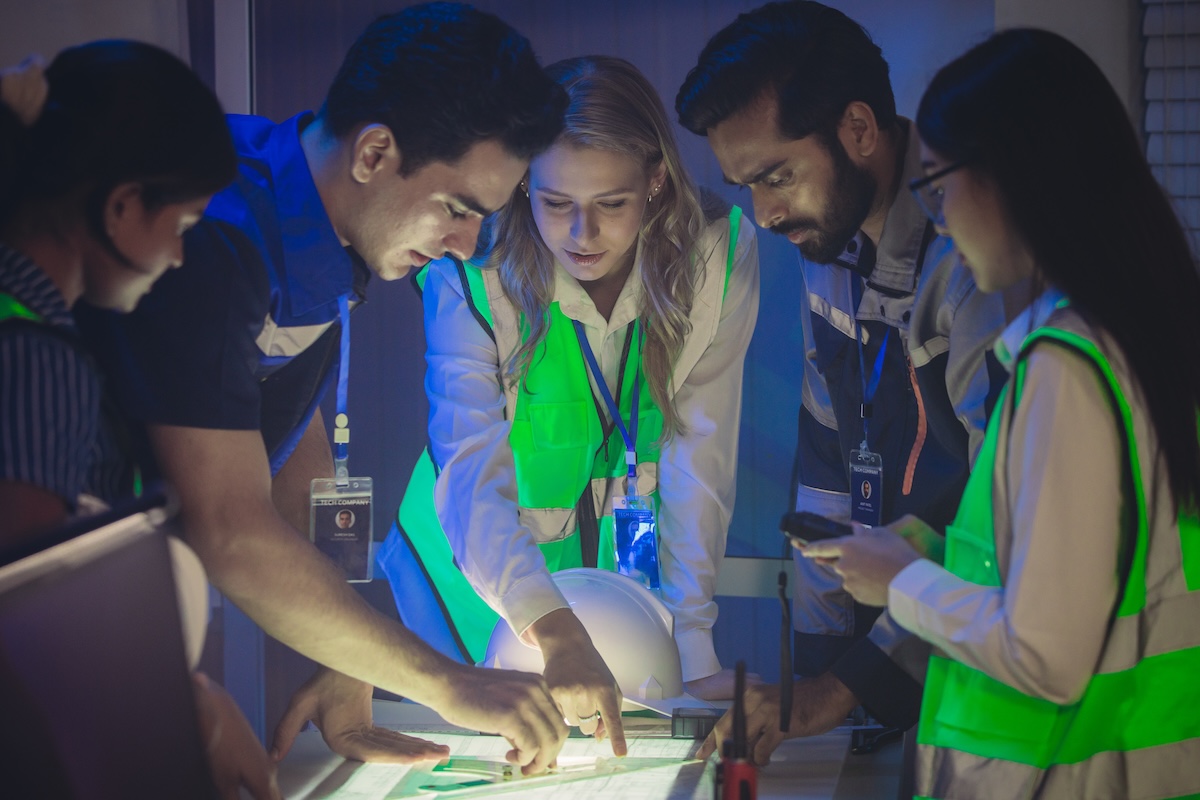 Engineers in high visibility vests collaborate in control room, reviewing digital site plans and dashboards during an operations and safety discussion. Coordination, monitoring, and incident readiness