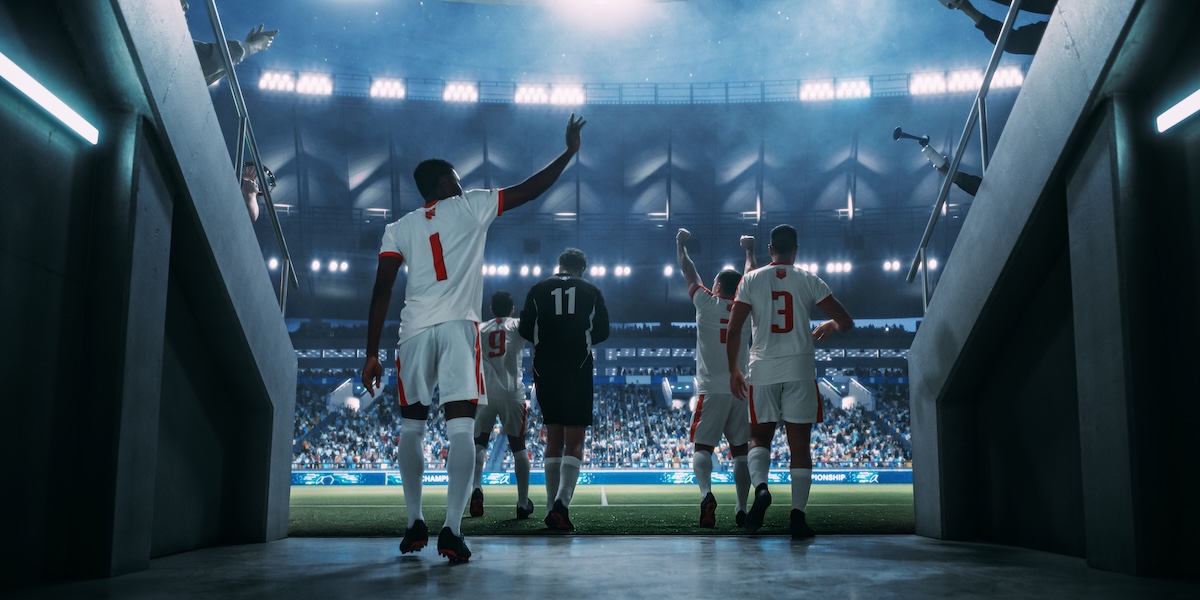 Male soccer players raise their hands to salute the crowds as they enter a sports arena.