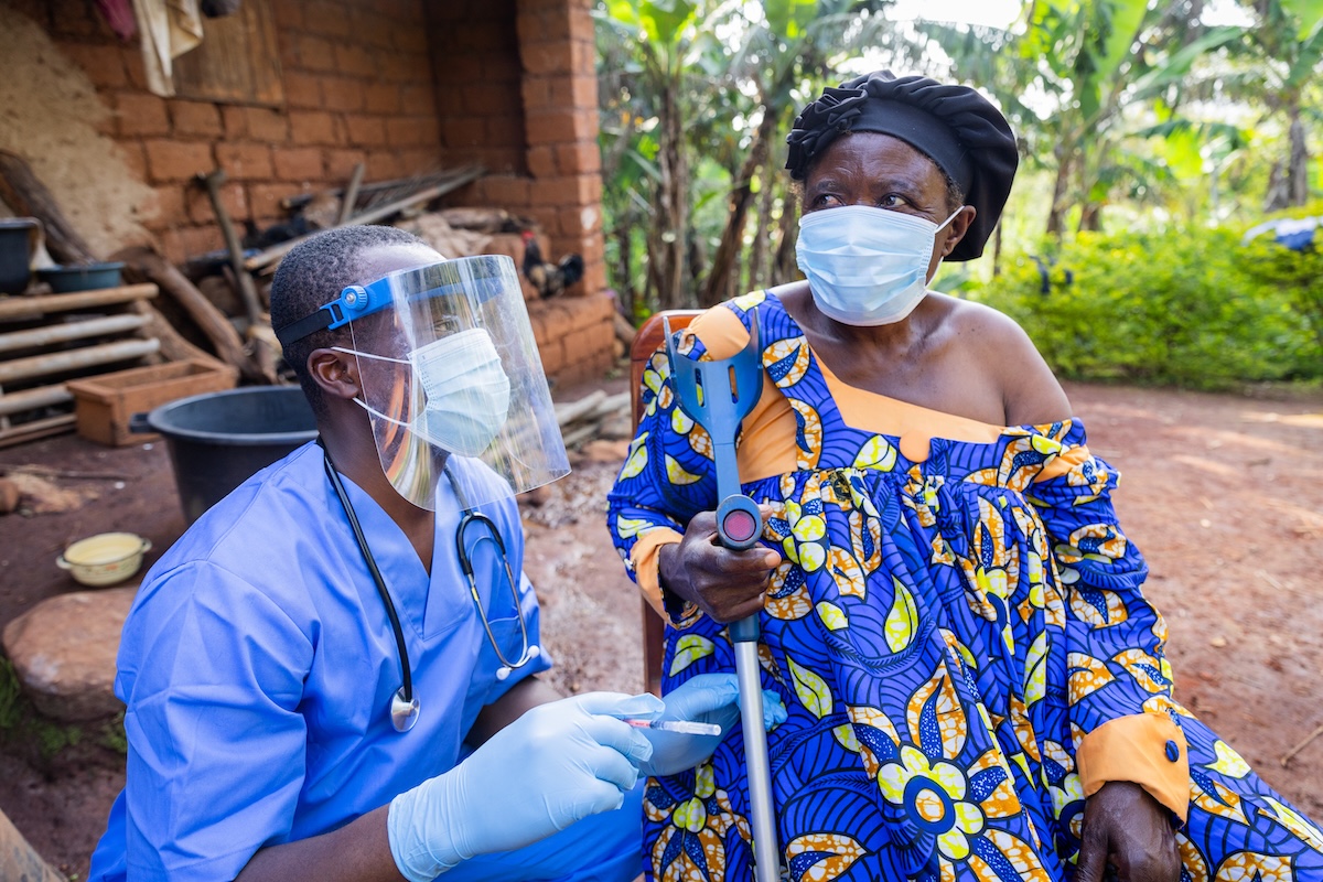 A male healthcare worker wears a face mask and crouches next to an old woman wearing a mask.